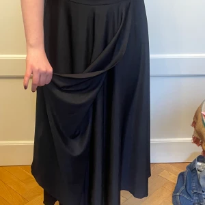 Vid silkig långkjol - Vid svart långkjol i tunt silkigt syntetmaterial med härligt fall och drapering fram. Midjemått 70 cm, längd 83 cm. Bred resår i midjan. Tvättas i 30 grader.