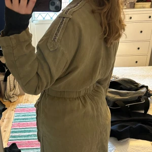 Grön vårjacka - Khaki jacka i 100% linne med detaljer av stenar. Knyts i midjan och går till mitten av låret ungefär. 