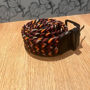 Flätat bälte  - Färg: Röd/orange/grå/svart/brun  120 cm långt men funkar för alla. Stretchigt tyg 