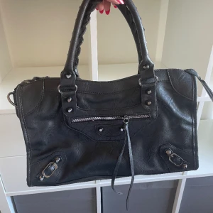 Svart väska - Oanvänd svart väska i fuskläder. Finns ett tillhörande längre band för att ha den över axeln