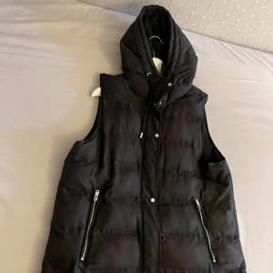 Svart väst med fickor  - Västen är bra i skick då den är använd under våren 2022-2023. Västen har ett lite hål som knappt syns.