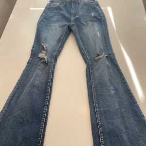Blå jeansbyxor med slitningar - Ett par blå jeansbyxor med slitningar på framsidan. Byxorna har en klassisk femficksdesign och är i en bootcut-stil.