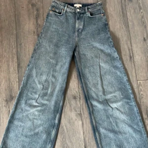 Blå jeansbyxor från H&M - Ett par blå jeansbyxor från H&M i storlek EUR 34. Byxorna har en hög midja och en vid passform. De är tillverkade i ett klassiskt jeansmaterial och har fickor både fram och bak.