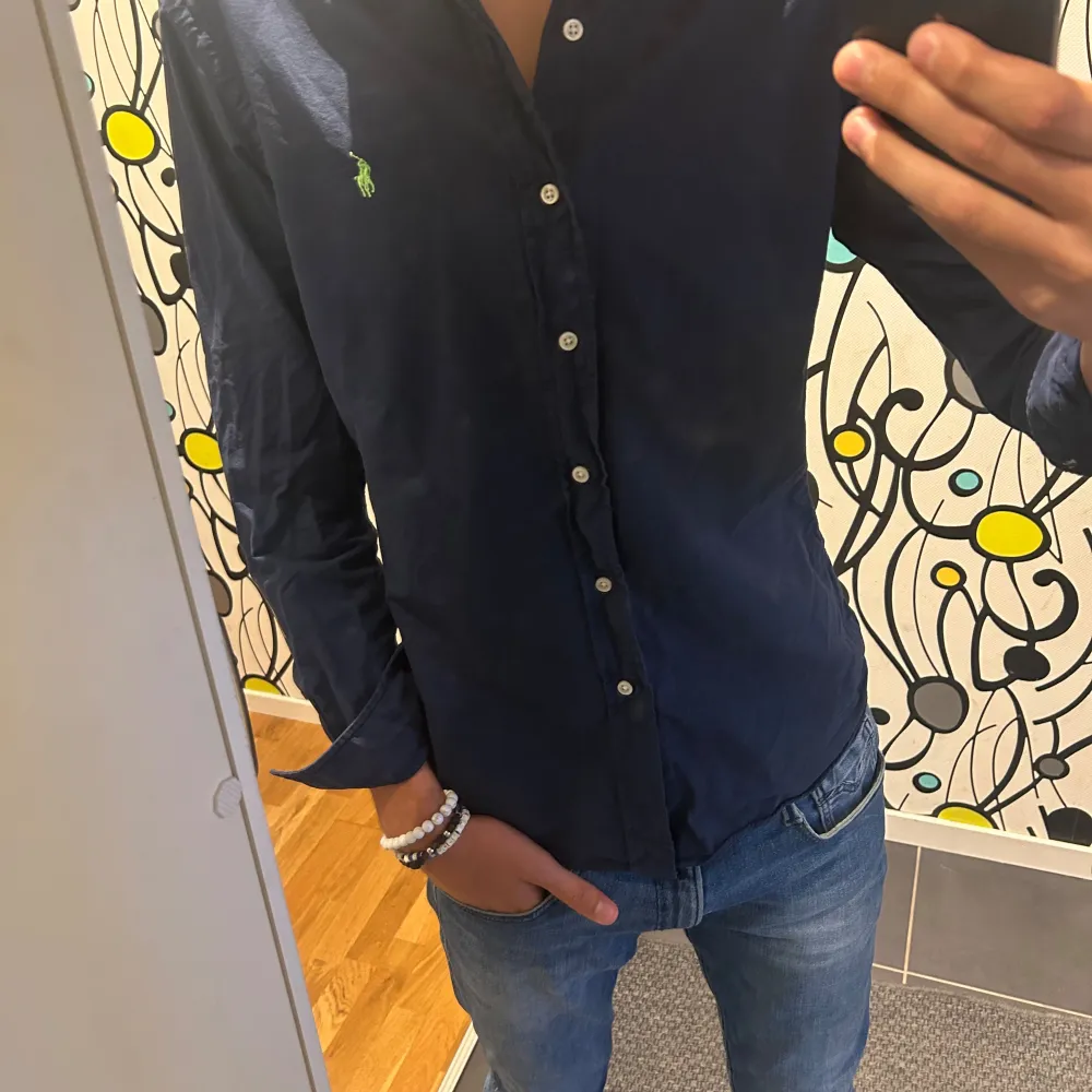 En mörkblå skjorta från Polo Ralph Lauren i storlek M. Skjortan har långa ärmar och knappar framtill. Den är av slim fit-modell och har en broderad grön logotyp på bröstet.. Skjortor.