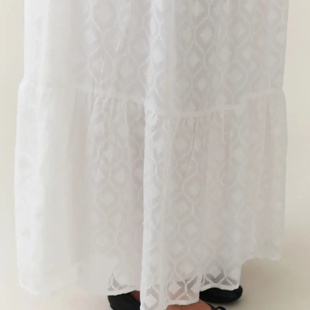 En elegant vit långkjol med ett subtilt mönster. Kjolen har en elastisk midja och är tillverkad i ett lätt och luftigt material, perfekt för varma dagar.. Hameet.