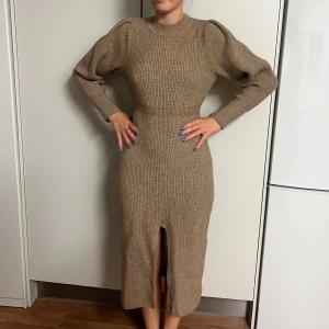 Beige stickad klänning från ADOORE - En elegant beige stickad klänning från ADOORE. Klänningen har långa ärmar och en slits framtill. Den är tillverkad av en blandning av akryl, polyamid, polyester, kashmir och nylon, vilket gör den både bekväm och varm.