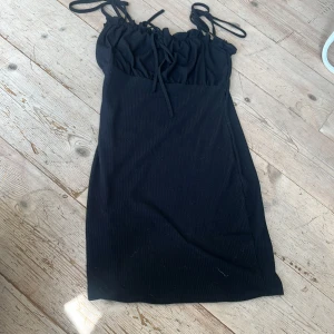 Svart klänning - Jag säljer nu denna svarta klänningen. Den är kort, har band som man knyter över axlarna och en rosett högst upp där fram. Den har även en söm över bröstet så att den blir lite puffigare där. Klänningen är i storlek XS. Stretchig, bra skick. 💗