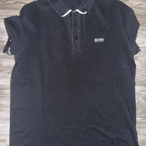 Svart BOSS pikétröja i storlek M - Säljer en svart pikétröja från BOSS i storlek M. Tröjan är i mycket bra skick och har en klassisk design med vita detaljer och BOSS-logo på bröstet samt ärmarna. Mjukt och bekvämt material.
