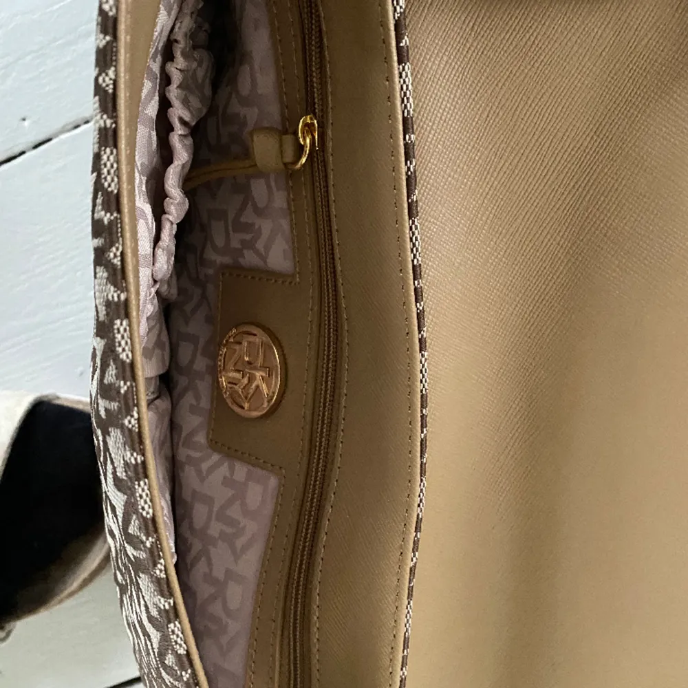 Helt ny DKNY handväska i brunt/beige skinn och beige tyg. Justerbar axelrem så väskan kan bäras cross body eller på ena axeln. Fack med dragkedja inne samt två mindre fack förutom större.  Svart dust bag medföljer. Nypris ca 1500 kr. . Väskor.