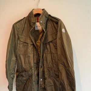 Moncler Bertrand Giubotto feeld jacket - Ofodrad jacka med dragkedja och knappar och huva i kragen. Olivgrön med Moncler stipes längs med dragkedjan.  Sparsamt använd och i mycket gott skick!  Storlek 1 (46-48 EU)