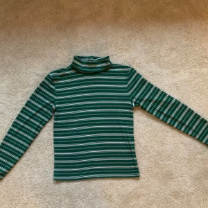 Grön, randig polotröja - I mycket gott skick. Lätt stretchigt tyg. Uppskattar storleken till en damstorlek 36/38. Mått när plagget ligger platt: axel till axel 41 cm; armhåla till armhåla 42 cm; axel till mudd 58 cm; totallängd (från axeln) 55 cm. Jag är 173 cm för referens.