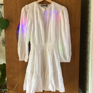 Vit klänning från Zara - En vit klänning från Zara med broderade detaljer och lång ärm. Klänningen har en smockad midja och är tillverkad i ett lätt och luftigt material. Den har en knappstängning vid halsen och är dekorerad med spetsdetaljer. Enbart använd ett fåtal ggr så i superskick 9/10. Skicka dm vid frågor.