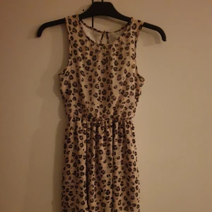 Leopardmönstrad klänning - En ärmlös klänning med leopardmönster. Klänningen har en beige basfärg med svarta och bruna fläckar. Den har en elastisk midja och en liten öppning med knapp i ryggen. Från H&M i stl 146