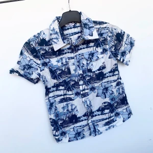 Skjorta barn  - Oanvänd kortärmad skjorta för barn i storlek 110/116. Vit med blåa blommor/palmer/skepp. Två stängbara bröstfickor. Vita knappar. 