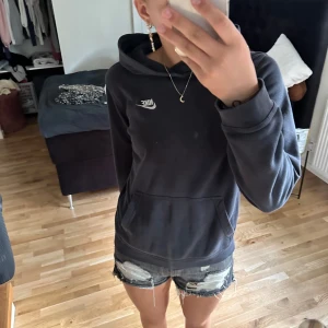 Svart hoodie från Nike - En svart hoodie från Nike med en vit logotyp på bröstet. Hoodien har en standard passform och en känguruficka framtill. Den är tillverkad i Kambodja och passar personer med en längd på 158-170 cm.