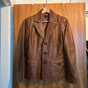 Brun skinnjacka från Lindex - En brun skinnjacka från Lindex i storlek 36. Jackan har långa ärmar, knappar framtill och två fickor med knappar. Den är tillverkad av 100% läder. Sparsamt använd. 