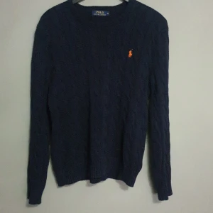 Mörkblå stickad tröja från Polo Ralph Lauren - En mörkblå stickad tröja från Polo Ralph Lauren med ett orange broderat märke på bröstet. Tröjan har långa ärmar och en rund halsringning. Den är helt nytt använd två gånger gillade inte den och vi kan snacka om priset.