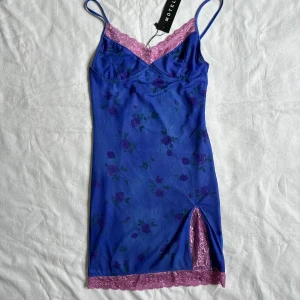 Blå klänning från Motel - helt ny klänning från Motel Rocks med blommigt mönster och rosa spetsdetaljer. Klänningen har smala axelband och en slits med rosa spetskant.