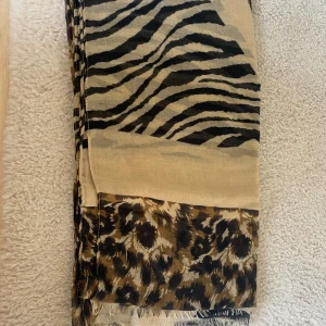 Beige och svart mönstrad halsduk - En beige halsduk med ett djurmönster i svart och brunt. Halsduken har både zebra- och leopardmönster, vilket ger den ett unikt och stilfullt utseende.