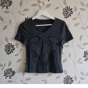 Grå t-shirt, kors, Strl: 36/S, wings, rhinestones, strass, y2k, 2000 - Grå t-shirt med kors, Strl: 36/S, wings, rhinestones, strass, y2k, 2000. Något croppad modell. OBS! Jag kan inte skicka något vecka 26.