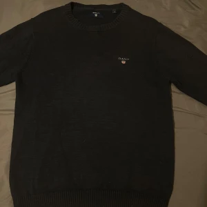 Svart stickad tröja från GANT - Säljer en klassisk svart stickad tröja från GANT med rund halsringning och broderad logga på bröstet. Tröjan har långa ärmar och ribbade muddar vid ärmslut och nederkant.