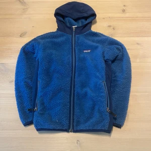 Blå fleecejacka från Patagonia - Mysig blå fleecejacka från Patagonia med huva och dragkedja framtill. Jackan har två fickor med dragkedja och förstärkta partier på armbågar och sidor. Skicka ett meddelande för frågor, storlek, mått osv. 
