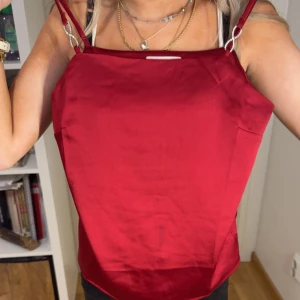 Röd satinlinne med smala axelband - Säljer ett rött linne i satin med smala axelband och rak halsringning. Perfekt för att styla med smycken och passar till många olika outfits. Linnet har en glansig finish och är lätt att bära.