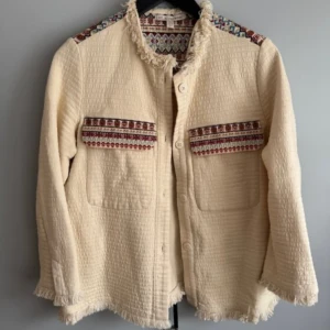 Beige jacka med mönstrade detaljer - Säljer en beige jacka med långa ärmar och dekorativa mönstrade band på axlar och fickor. Jackan har knappar framtill och råa kanter vid ärmslut och krage. Perfekt för dig som gillar unika detaljer och boho-stil.