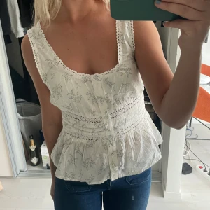 Vit blommig blus med spetsdetaljer - Söt vit blus med blommigt mönster och spetsdetaljer längs kanterna. Blusen har knappar framtill, markerad midja och volang nertill. Perfekt för en somrig look! Aldrig använd! 