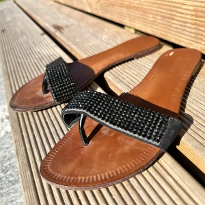 Svarta sandaler med glittriga detaljer  - Säljer ett par snygga sandaler i brunt och svart med breda remmar täckta av svarta glittriga stenar. Perfekta för sommaren och lätta att matcha till olika outfits.