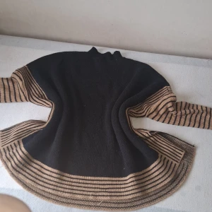 Svart och beige stickad tröja med randiga detaljer - Säljer en svart stickad tröja med beige och svarta ränder längs ärmar och nederkant. Tröjan har lång ärm och hög hals, perfekt för kyligare dagar. Mjuk och skön med snygg kontrast mellan färgerna.