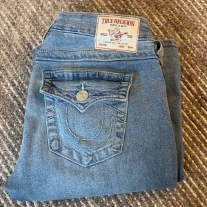 Blå bootcut jeans från True Religion - Säljer ett par klassiska blå jeans från True Religion med bootcut passform. Jeansen har en snygg fickdetalj med knapp bak och tydliga sömmar. Perfekta för dig som gillar en tidlös denimlook.