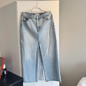 Ljusblå jeanskjol från Stradivarius - Lång ljusblå jeanskjol från Stradivarius med klassisk femficksdesign och hög midja. Kjolen har en tydlig slits framtill som ger en snygg detalj och avslappnad känsla. Perfekt att styla med sneakers eller boots.