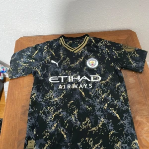 Manchester City fotbollströja Treble Winners - Snygg Manchester City fotbollströja från Puma med svart bas och guld- samt grått mönster. Tröjan har klubbmärke på bröstet, Treble Winners-detaljer och Etihad Airways sponsortryck. Perfekt för fans som vill sticka ut på läktaren.