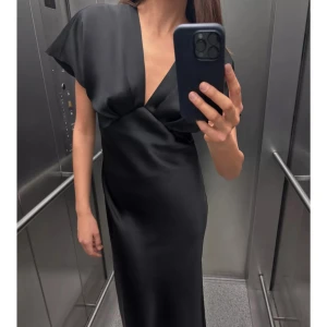 Svart långklänning i satin med v-ringning - Elegant svart långklänning i satin med djup v-ringning och korta, vida ärmar. Klänningen har en omlottliknande överdel. Helt ny från Zara, med prislappen kvar. 