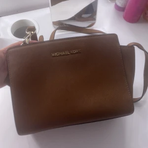 Brun axelväska från Michael Kors - Säljer en snygg brun axelväska från Michael Kors med guldfärgade detaljer och logga framtill. Köpte för 4000kr fast säljer nu för 1000 eller lite mindre för att den är lite sliten. Väskan har justerbar axelrem och flera fack inuti för bra organisering. Perfekt för att bära med sig det viktigaste i vardagen.