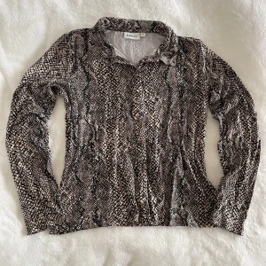 Ormmönstrad skjorta från Kappahl - Säljer en långärmad skjorta från Kappahl med ormmönster i svart, beige och grått. Skjortan har klassisk krage och knäppning framtill. Perfekt för dig som vill sticka ut med ett djurmönster.