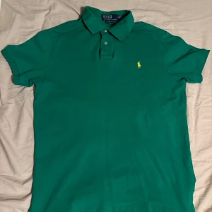 Grön pikétröja från Polo Ralph Lauren - Säljer en klassisk grön pikétröja från Polo Ralph Lauren med gul broderad logga på bröstet. Tröjan har krage, knappar och korta ärmar. Perfekt för en stilren och sportig look.