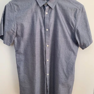 Blå kortärmad skjorta från Selected Homme - Snygg blå skjorta från Selected Homme i modellen Identity slim fit. Skjortan har korta ärmar, klassisk krage och knäppning framtill med vita knappar. Perfekt för en stilren och enkel look.