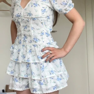 Vit blommig kortklänning med volanger - Supersöt vit kortklänning med blått blommönster och flera volanglager. Den är lite insydd i midjan men väldigt lite. Använd en gång på midsommar.  Den är storlek S och vet inte vart den kommer ifrån. Nypris 600