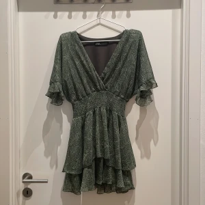 Grön mönstrad kortklänning från ZARA - Supersöt grön kortklänning från ZARA med abstrakt mönster, v-ringning och volangdetaljer. Klänningen har korta ärmar med volang och smockad midja för en smickrande passform. Mycket fint skick, använd enstaka gånger. Kameran kan inte riktigt plocka upp färgen korrekt, klänningen är mer grön i verkligheten! Går inte att köpa på zara längre dessutom.