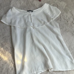 Vit blus med volang - Säljer en söt vit blus med volangdetalj längs halsringningen. Blusen har en lös passform och är tillverkad i ett lätt, strukturerat tyg. Perfekt för vår och sommar! Köpt i spanien lite osäker på storlek med sitter bra på mig som vanligtvis har xs-s i blusar💞