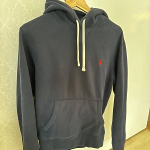 Mörkblå hoodie från Ralph Lauren - Mörkblå hoodie från Ralph Lauren med klassisk känguruficka och vit dragsko i huvan. Tröjan har en röd broderad logga på bröstet och är långärmad. Perfekt för en avslappnad stil.