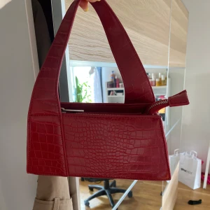 Röd axelväska i krokopräglat mönster - Säljer en snygg röd axelväska med krokopräglat mönster och bred axelrem. Väskan har dragkedja upptill och en stilren, boxig form. Perfekt accessoar för att lyfta din outfit.
