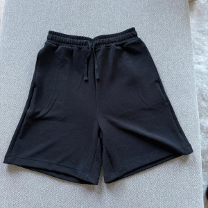 Svarta shorts med snören - Svarta shorts med elastisk midja och snören. Modellen har en avslappnad passform och är tillverkad i ett mjukt material som passar perfekt för vardagsbruk eller träning. Två sidofickor för enkel förvaring. Perfekt sommarplagg