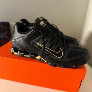 Svarta Nike sneakers med guld-detaljer - Säljer ett par svarta Nike sneakers med guldiga detaljer och logga. Skorna har snörning, platt sula och en sportig design med mesh och syntetmaterial. Perfekta för dig som vill sticka ut med stilrena och unika sneakers.