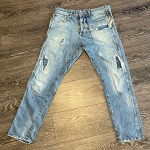 Jack&jones Erik Anti fit  - Säljer ett par blå jeans med flera slitningar och hål på benen. Jeansen har klassisk femficksdesign och knappgylf. De har en rak passform och är perfekta för en avslappnad streetstil. Erik Anti fit. 
