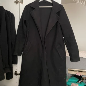 Svart kappa  - Säljer en svart kappa med bred krage och långa ärmar. Jackan har två snedställda fickor framtill och en klassisk, stilren design. Perfekt för kyligare dagar och enkel att matcha med olika outfits. 