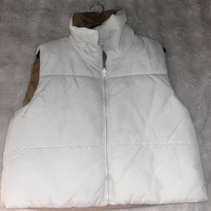 Vändbar vit och beige dunväst - Säljer en vändbar dunväst i vitt och beige med hög krage och dragkedja framtill. Västen är vadderad och har en enkel, stilren design utan synliga fickor eller mönster. Perfekt för lager-på-lager under kyliga dagar.