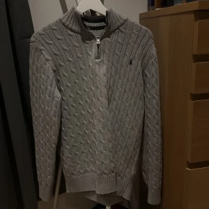 Grå kabelstickad tröja från Polo Ralph Lauren - Säljer en grå kabelstickad tröja från Polo Ralph Lauren med halv dragkedja och hög krage. Tröjan har lång ärm, ribbade muddar och en liten broderad logga på bröstet. Perfekt för kyliga dagar. OBS! Den är XL i barnstorlek inte vuxen